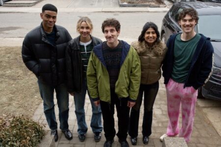 Five young adults smiling and standing outdoors, dressed in casual winter clothing.