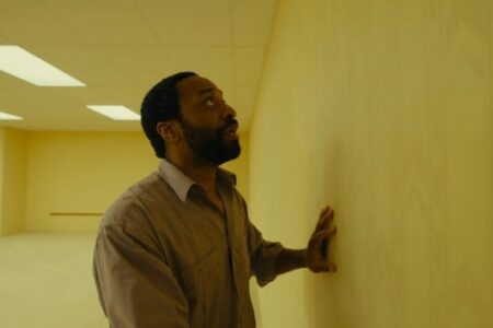 Bearded man in a beige shirt touches a pale yellow wall in an empty, fluorescent-lit room, looking up.