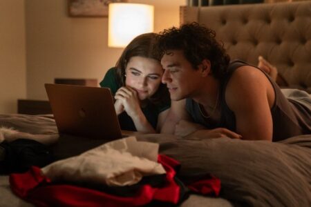 Two young adults lie on a bed, illuminated by a laptop screen, watching content together in a dimly lit room.