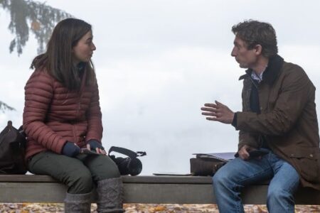 Two people, a woman in a red jacket and a man in a brown jacket, sit on a bench outdoors, engaged in conversation.