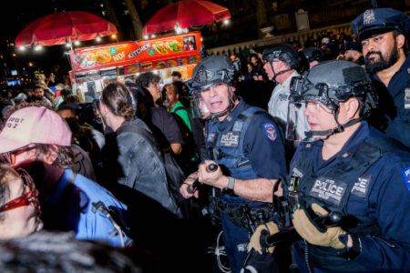 Police in tactical gear face a large crowd at night after a Knicks game, with a food cart in the background.