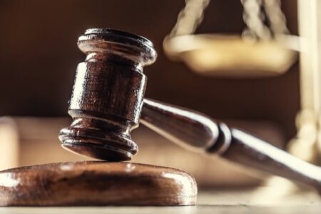 A wooden judge's gavel rests on a sound block, with blurred scales of justice in the background, symbolizing law.