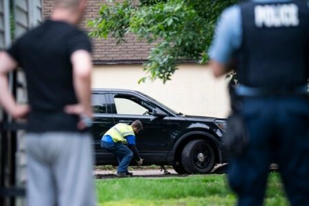 A person in a safety vest works near a black SUV, observed by a police officer and another person.