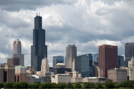 The Chicago skyline featuring the iconic Willis Tower and numerous other skyscrapers under a cloudy sky, with trees in the foreground.