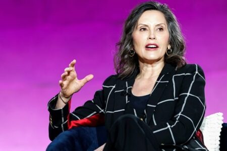 Michigan Governor Gretchen Whitmer speaks, gesturing with her right hand against a purple background.