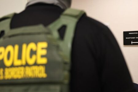 Back of a US Border Patrol officer in a tactical vest, walking past an 'Immigration Court' sign.