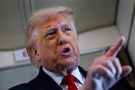 Donald Trump speaking and pointing, wearing a dark suit and red tie, with a serious expression.