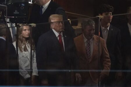 Donald Trump in a suit and red tie, with others, watches a Knicks game from a box at Madison Square Garden.