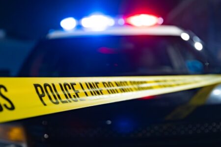 Police car with flashing red and blue lights behind yellow 'POLICE LINE DO NOT CROSS' tape at night.