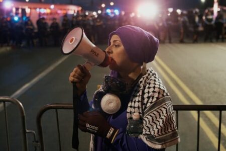A woman in a purple head covering and keffiyeh speaks into a megaphone at a night protest, with police lights behind her.