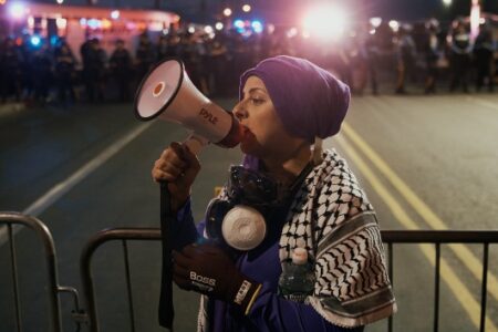Woman in purple headscarf and keffiyeh speaks into megaphone at nighttime protest with police lights in background.