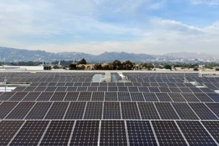 A vast array of solar panels covers a rooftop with a city and mountains visible in the background under a partly cloudy sky.