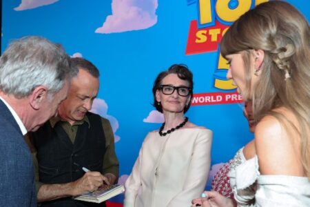 Tom Hanks signs a 'Toy Story' VHS tape for Taylor Swift at an event, with other attendees nearby.