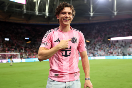 Tom Holland smiles, pointing to his pink Inter Miami CF soccer jersey on a field with stadium lights in the background.