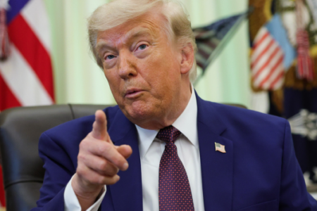 Donald Trump, in a suit and tie, points his finger while seated in the Oval Office.
