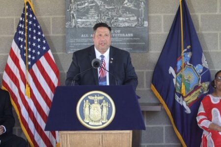 Assemblyman Brian Manktelow speaks at a podium with the New York State seal, flanked by American and NY flags.