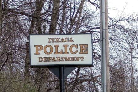 A white sign with brown lettering reads 'Ithaca Police Department' against a backdrop of bare trees.