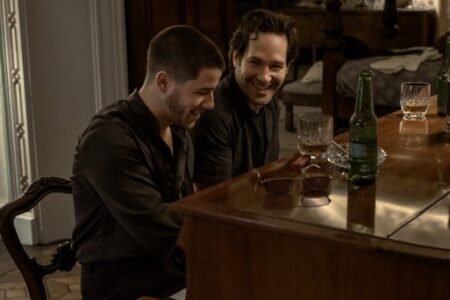Nick Jonas and Paul Rudd smiling and laughing together at a table with drinks.