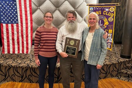 Malcolm MacKenzie, a longtime educator, holds an award plaque with two women, an American flag, and a Lions Club banner.