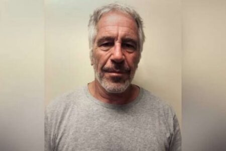 Jeffrey Epstein, a man with gray hair and beard, wearing a gray t-shirt, looks directly at the camera.