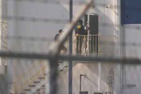 Federal agents on a balcony and stairs of an industrial building, seen through a fence, during a search warrant.