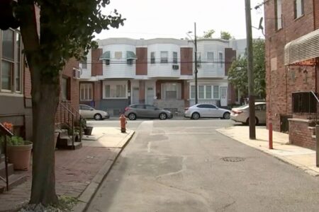 A quiet residential street in Philadelphia lined with brick row houses and parked cars under a clear sky.