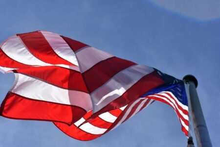 An American flag waves prominently against a clear blue sky, with its red and white stripes and blue field visible.