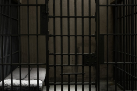 An empty, dark jail cell with metal bars in the foreground and a bed with a white mattress.