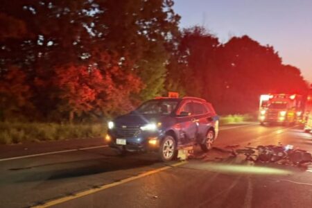 Accident scene with a damaged blue SUV, motorcycle debris on the road, and an emergency vehicle with flashing lights.