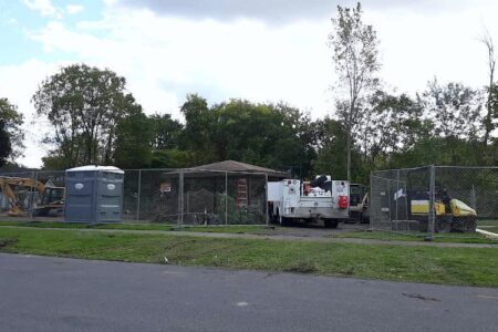 Construction site of Wall St Playground renovations in Auburn, NY, with heavy equipment and a portable toilet behind a fence.