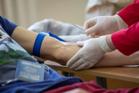 A medical professional prepares to draw blood from a donor's arm, with a blue tourniquet visible.