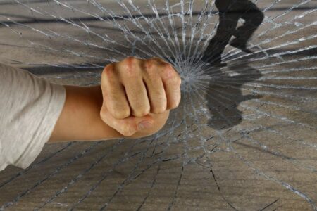 A fist punches through shattered glass, with a blurry figure running in the background, symbolizing vandalism or a break-in.
