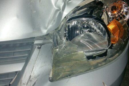 Close-up of a silver car's front with a severely damaged headlight and crumpled fender after a crash.
