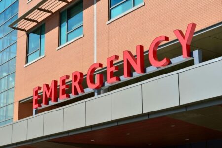 Close-up of a red 'EMERGENCY' sign on the brick and glass facade of a hospital or medical building.