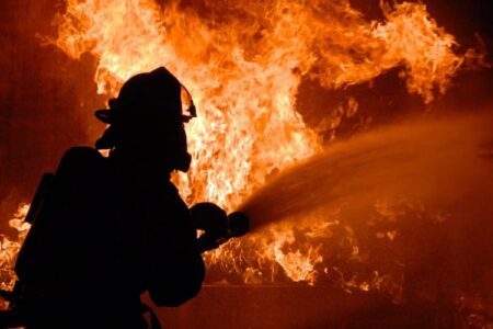 Silhouette of a firefighter spraying water into a large, intense fire.