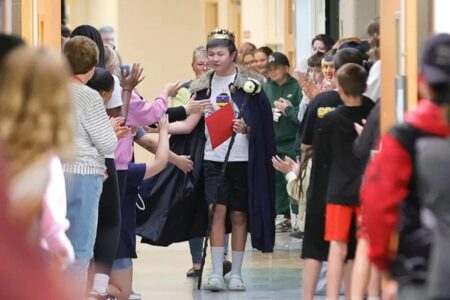 A student wearing a crown and cape walks through a school hallway, high-fiving classmates celebrating his spelling bee achievement.