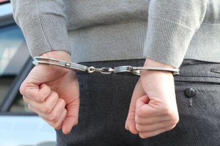 Close-up of a person's hands handcuffed behind their back, wearing a grey sweater and dark pants.