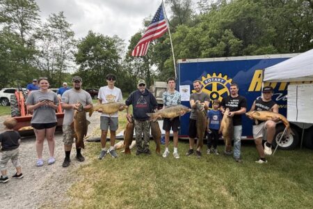 Participants proudly display their large carp catches at the Waterloo Carp Derby, with a Rotary trailer and American flag.