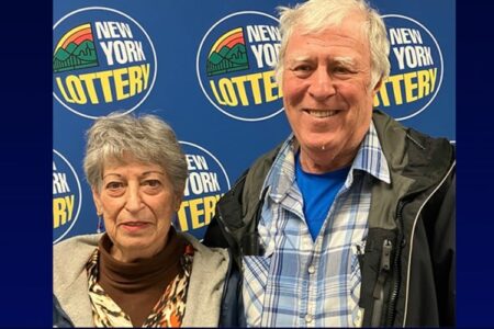 An older couple smiles in front of a New York Lottery backdrop after claiming their jackpot.