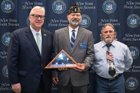 Three men, including a veteran holding a folded American flag, pose at a New York State Senate induction ceremony.