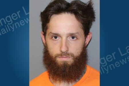 Mugshot of a man with a beard and brown hair, wearing an orange shirt.
