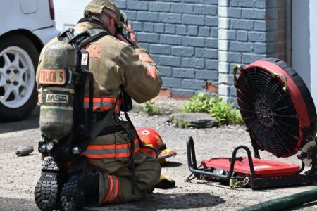A Syracuse firefighter kneels next to a ventilation fan, adjusting their gear after responding to a fire.