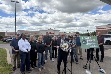 Senator Chuck Schumer speaks outside a Tops grocery store, holding a Senate seal, with a sign for 'Lower Prices' and a crowd.