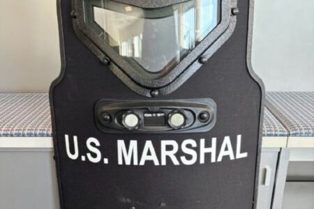A black ballistic shield with 'U.S. MARSHAL' written in white letters on its front.