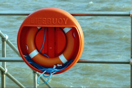 An orange lifebuoy in a holder attached to a metal railing, with rippling water visible in the background.