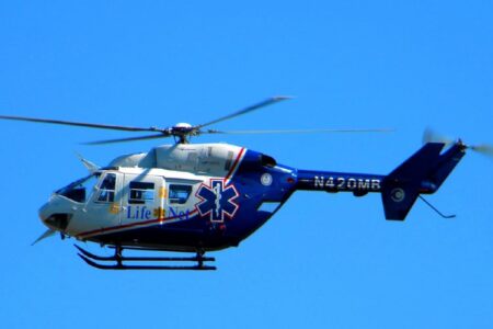 A LifeNet medical helicopter, blue and white with the Star of Life, flies against a clear blue sky, used to airlift a crash victim.