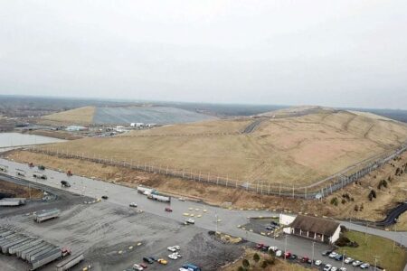 Aerial view of a large landfill with mounds of earth, roads, trucks, and a building under an overcast sky.