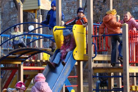Children play on a colorful playground structure, climbing, sliding, and standing on platforms.