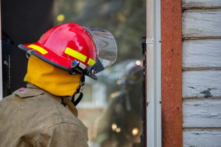 A firefighter in a red helmet and yellow hood looks into a window, with a reflection visible.