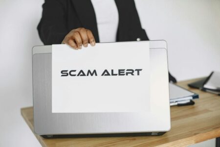 A person holds a 'SCAM ALERT' sign in front of a laptop, warning about potential fraud.